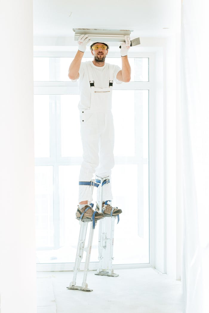 hero-01 A construction worker in white jumpsuit on stilts plastering a ceiling indoors.