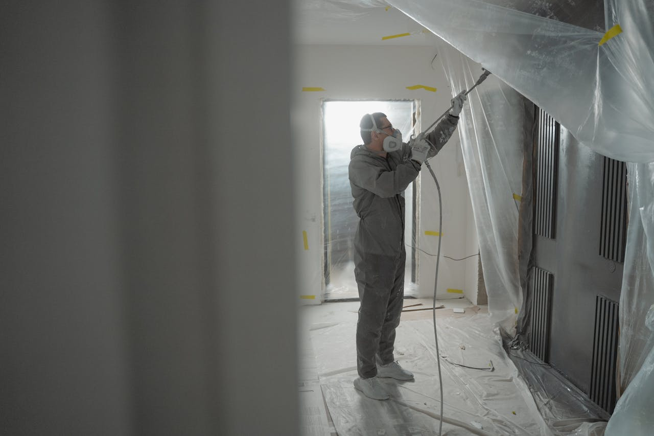 services-02 Man in protective gear spray painting a house's interior during renovation work.